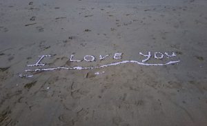 Image shows I Love You written in the sand on the beach