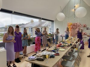 Image shows a room with people standing around a long table with food on it. There is a wide panoramic window on one wall of the room.