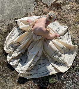 Image shows a woman seen from above, wearing a strapless white dress with a full skirt, she is sitting on a rock on the beach.