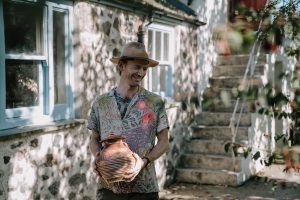Image shows a white male wearing a hat, and holding a ceramic pot, outside of a stone building