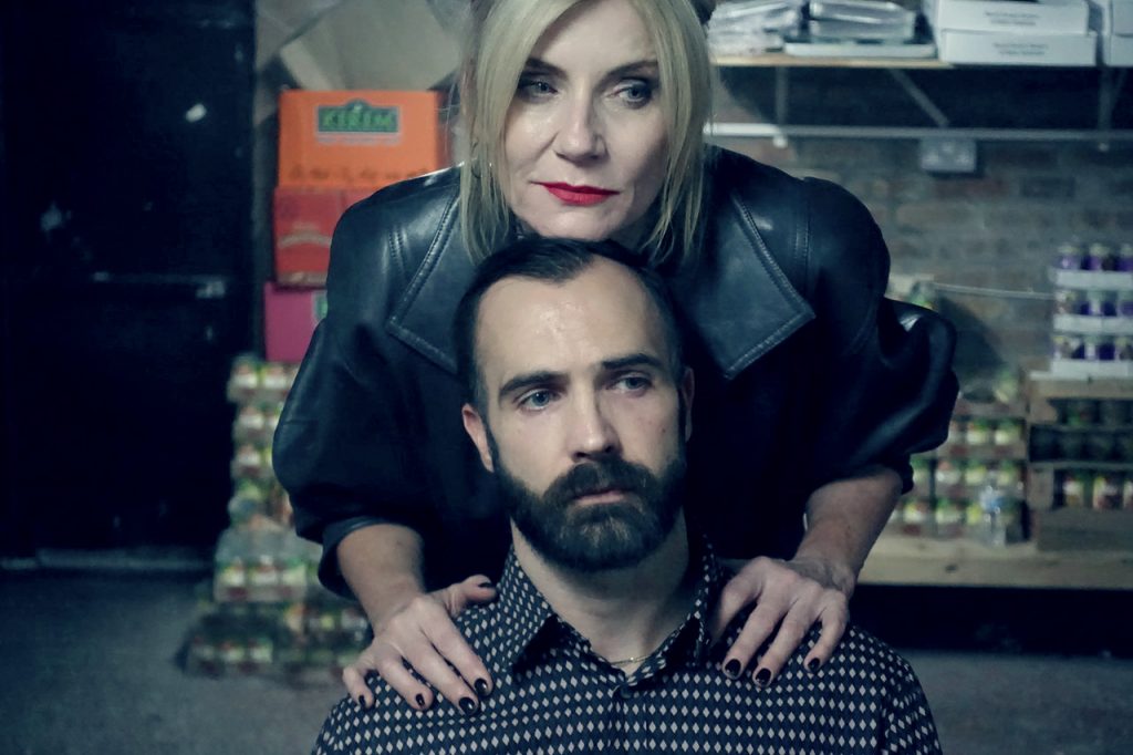 Image shows a white woman with blond hair standing with her nhands on the shoulders of a white man with dark hair and beard