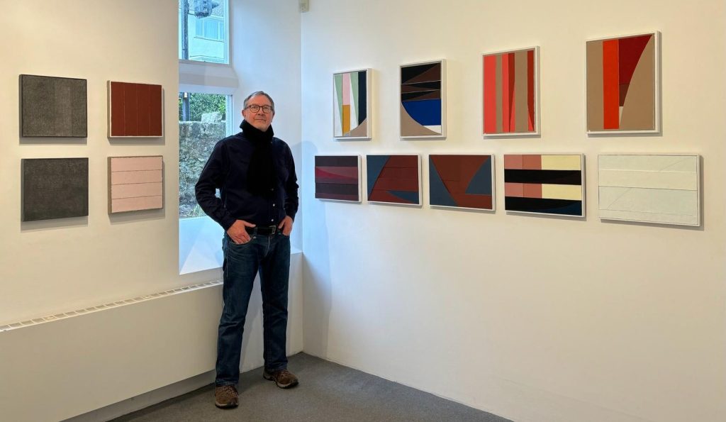 Image shows a white man in a drak top standing in corner with a window behind him and paintings on the wall to his right