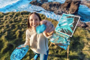 Image shows a white woman standing oustide on a bright day, in front of an easel which is located on the cliffs