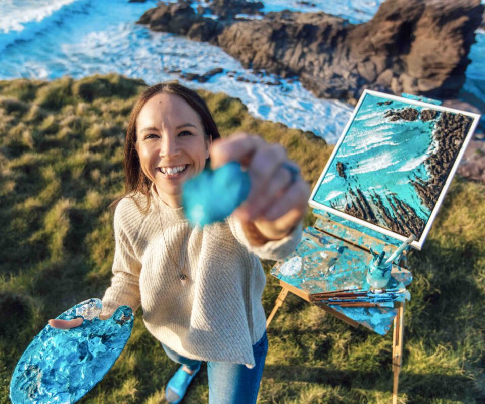 Image shows a white woman standing oustide on a bright day, in front of an easel which is located on the cliffs