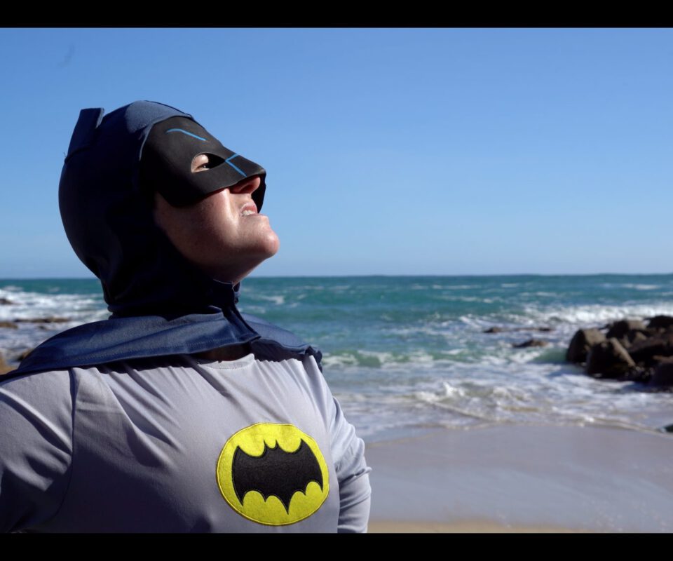 Person wearing a batman costume and hood stood on a sandy beach with the sea behind them,