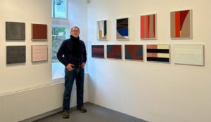 Image shows a white man in a drak top standing in corner with a window behind him and paintings on the wall to his right