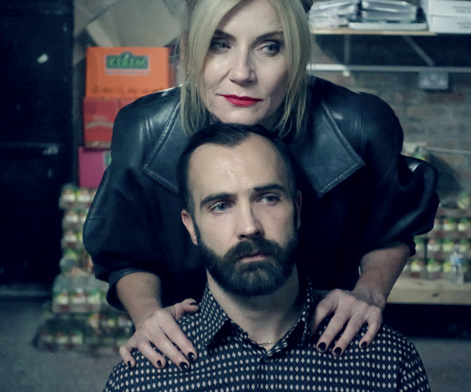 Image shows a white woman with blond hair standing with her nhands on the shoulders of a white man with dark hair and beard