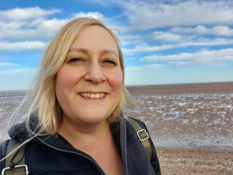 Image shows a white woman with blonde hair standing on a sandy beach with her back to the sea