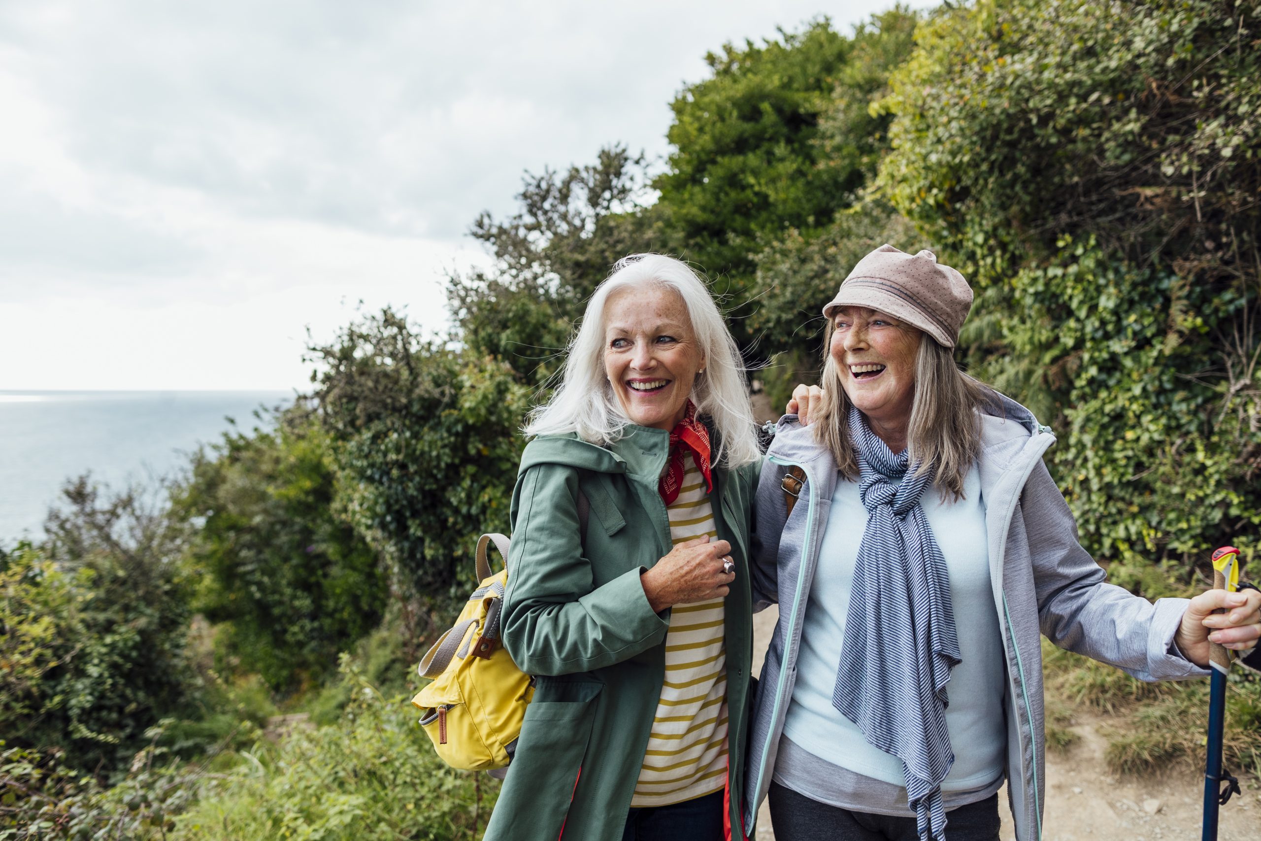 Female friends on a hike in Cornwall, they are walking on a coastal path, laughing while they have their arms around each other and looking at the view. The sea can be seen in the distance.