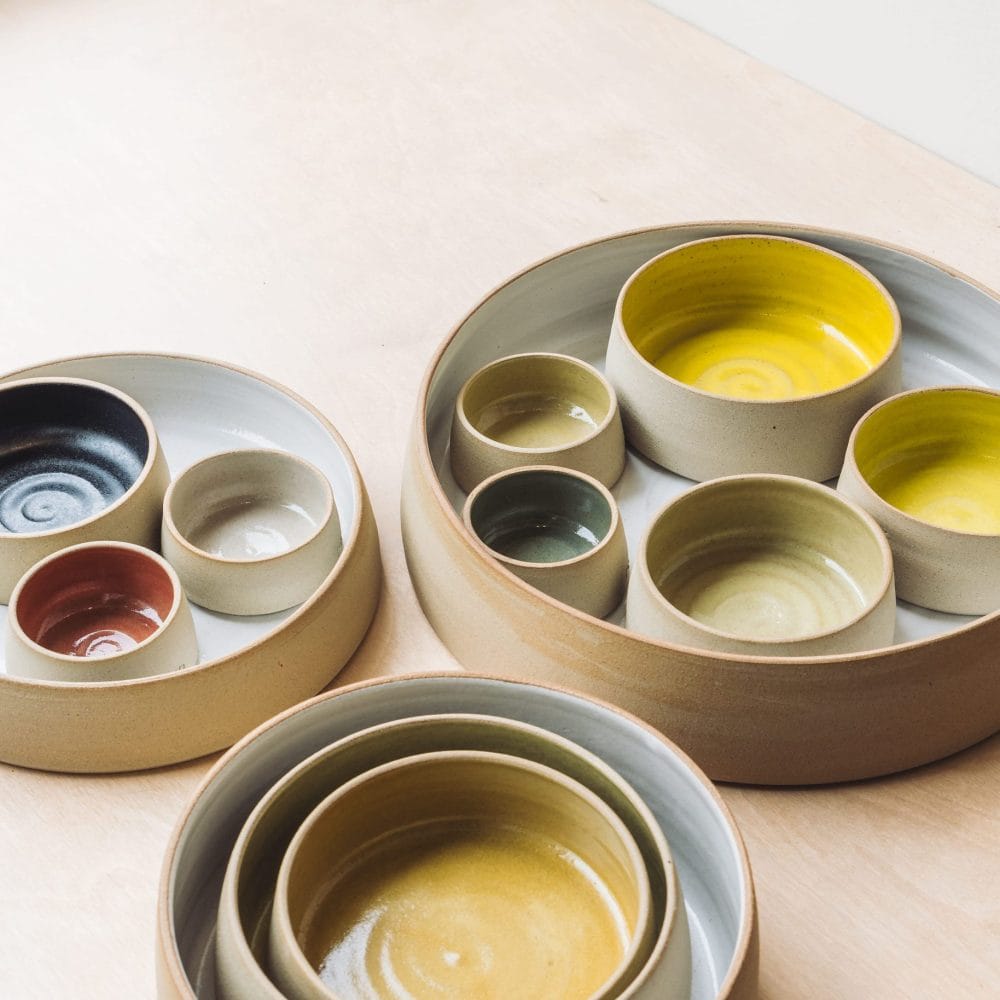 A set of round ceramic bowls in various sizes and earthy tones—yellow, green, black, and brown—nest neatly on a light wooden surface, from a mixed media group exhibition.