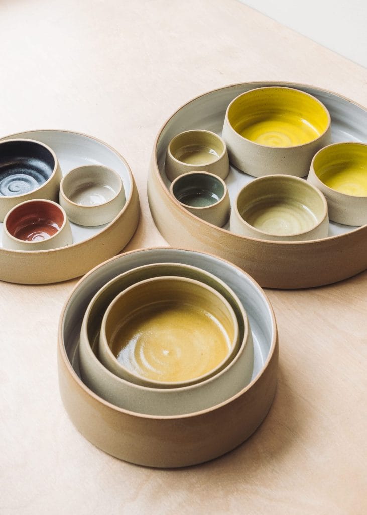 A set of round ceramic bowls in various sizes and earthy tones—yellow, green, black, and brown—nest neatly on a light wooden surface, reminiscent of pieces from a mixed media group exhibition.