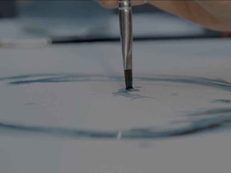A hand holds a paintbrush and paints a blue circular shape on white paper, creating a watercolour effect that highlights disability justice in visual arts.