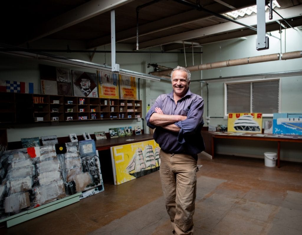 A man with grey hair and glasses stands smiling with arms crossed in a spacious art studio, surrounded by colourful paintings and artwork displayed on shelves and tables.