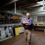 A man smiles with arms crossed in an art studio filled with colourful paintings and supplies. The vibrant space, lit by overhead lights, captures the creative spirit of the work around him.