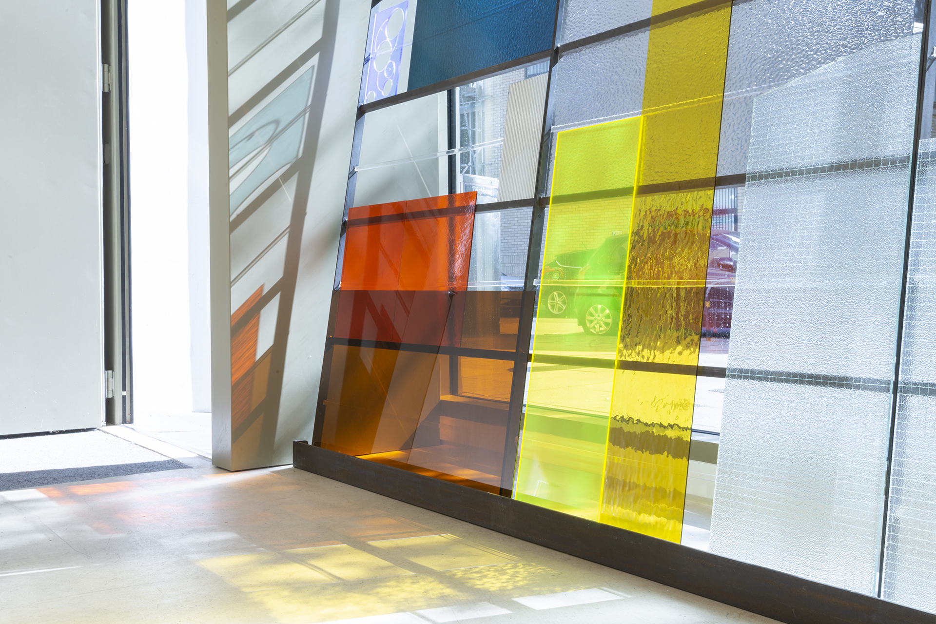 Colourful glass window panel in shades of orange, yellow, blue, and clear by Abigail Reynolds, sunlight casts vibrant geometric shadows across the bright, contemporary interior space.