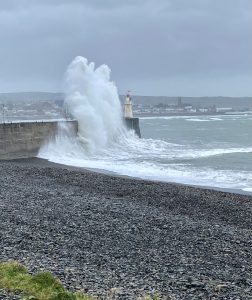 Image shows large wave crashing over Newlyn pier and lighthouse