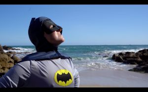 Person wearing a batman costume and hood stood on a sandy beach with the sea behind them,