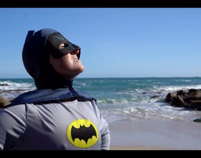 Person wearing a batman costume and hood stood on a sandy beach with the sea behind them,