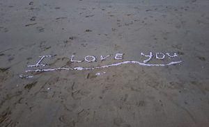 Image shows I Love You written in the sand on the beach