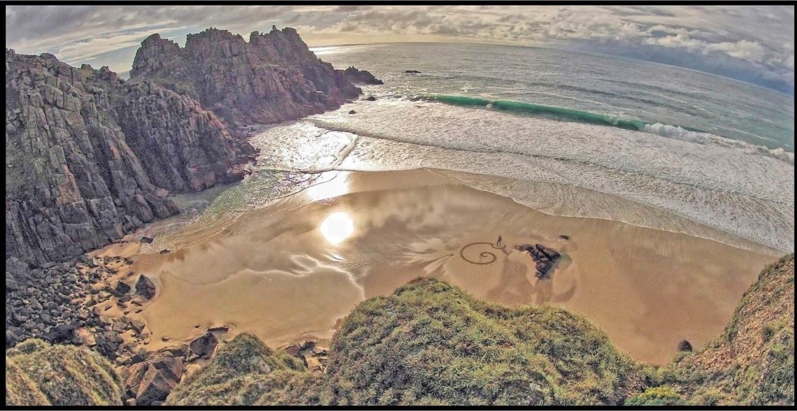 Looking down on a sandy beach where a small figure is creating patterns in the sand