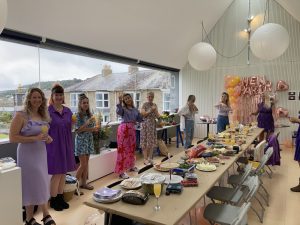 Image shows a room with people standing around a long table with food on it. There is a wide panoramic window on one wall of the room.