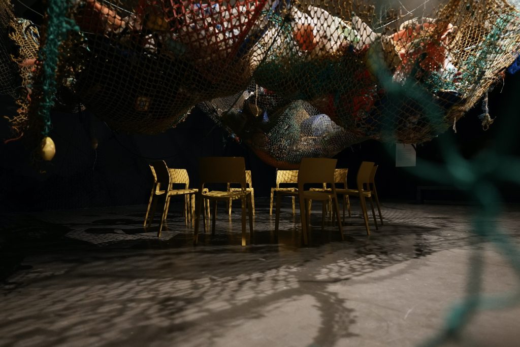 Image shows a large fishing net suspended in a gallery space, and full of marine rubbish with yellow chairs below the net