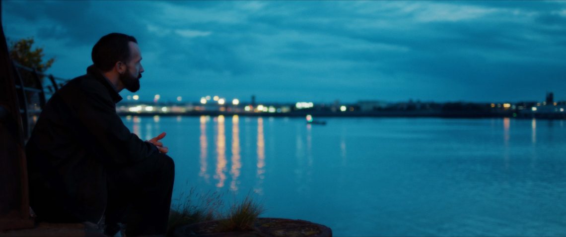 Image shows a wide shot of a river at night with a man sitting on the bank