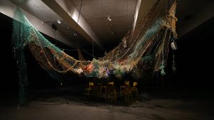 Image shows a large fishing net suspended in a gallery space, and full of marine rubbish