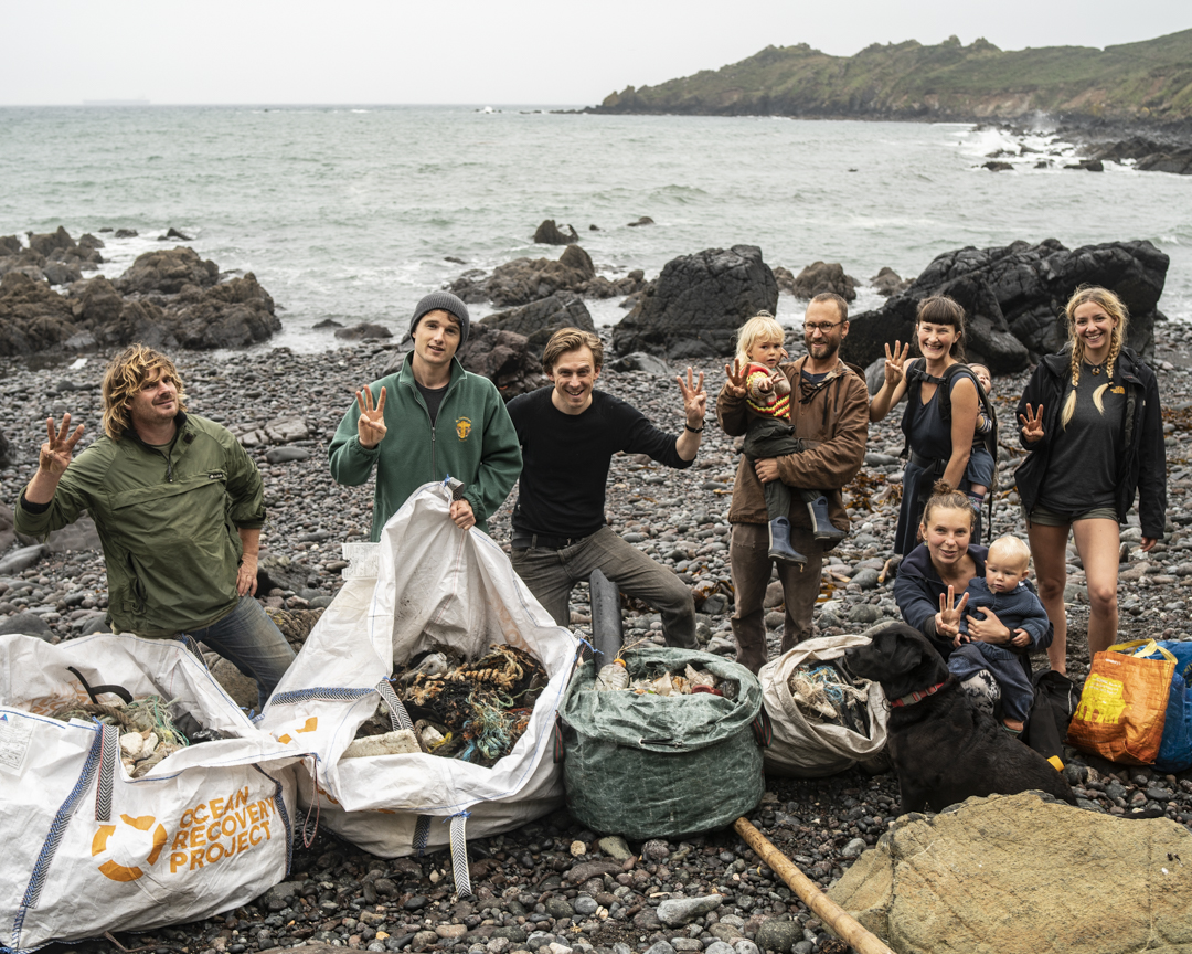 Image shows a group of people standing on rocky beacj with large bags of marine rubbish in front of them