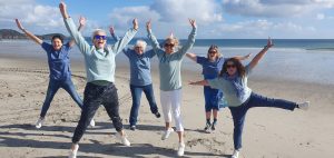 Image shows five wite women wearing jeans and sweatshirs, jumping up on a sandy beach