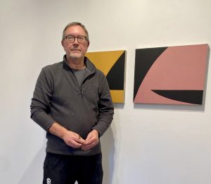 Image shows a white man wearing a grey top standing in front of two abstract paintings on the wall